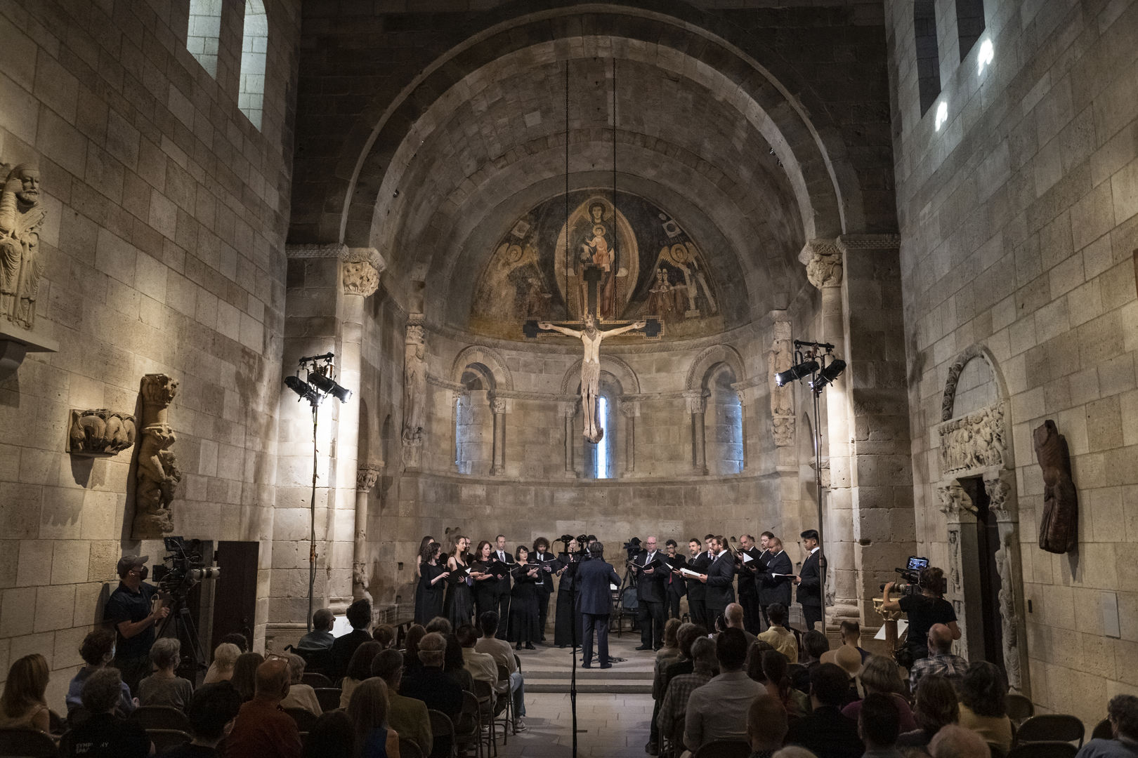 A choir sings underneath a 12th-century apse and Jesus figure. An audience watches.